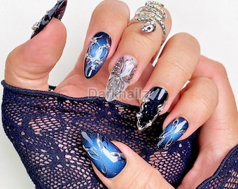 Peut inclure: Une main présente une manucure avec des ongles artificiels longs et ovales. Les ongles sont bleu foncé avec des accents argentés et blancs. Certains ongles ont un effet dégradé, tandis que d'autres présentent des motifs et des embellissements complexes. Une bague serpent argentée est également visible.