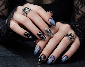 Puede incluir: Primer plano de manos con uñas largas, azul oscuro y negras, adornadas con intrincados diseños plateados. Anillos plateados, uno con un diseño de calavera, adornan los dedos. El fondo oscuro resalta el arte de uñas y las joyas.
