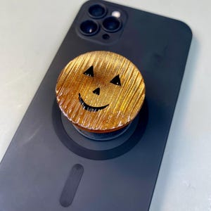 May include: A circular phone grip featuring a metallic gold, textured finish with a carved jack-o'-lantern face. The design includes two triangular eyes, a small triangular nose, and a curved mouth, all in black, attached to a matte black smartphone.