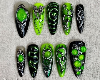 Op de afbeelding: Tien lange, groene en zwarte nageltips met zilveren accenten. De tips hebben verschillende ontwerpen, waaronder een vlinder, wijnstokken, swirls en een slang. Sommige tips hebben groene glitteraccenten.