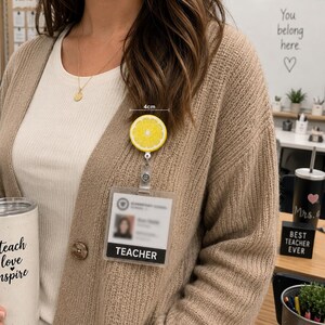 May include: A yellow lemon-shaped badge reel, approximately 4cm in diameter, worn on a beige cardigan. The badge reel is attached to a clear ID holder with the word "TEACHER" printed on it. A white tumbler with the words "teach love inspire" is also visible.
