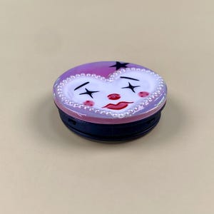 Peut inclure: Un support de t&eacute;l&eacute;phone circulaire avec une surface violette brillante et un motif de visage en forme de c&oelig;ur blanc. Le visage pr&eacute;sente des yeux en forme d'&eacute;toile noire, un nez rouge, des l&egrave;vres rouges et des joues roses. Le c&oelig;ur est bord&eacute; de perles blanches.