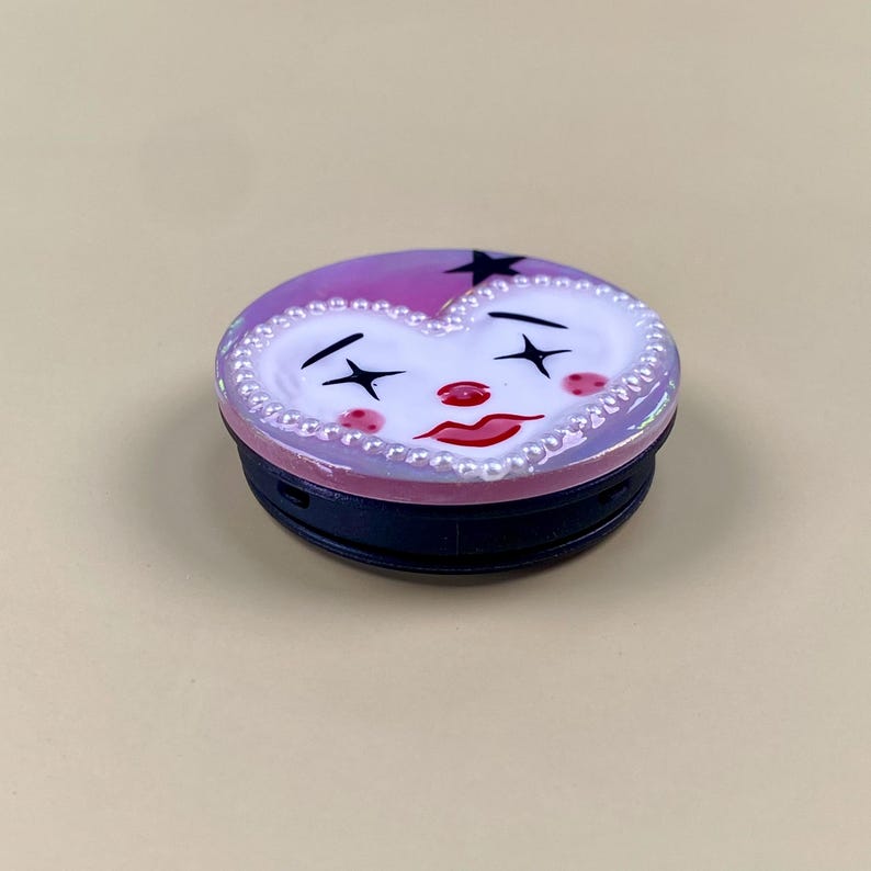 Peut inclure: Un support de t&eacute;l&eacute;phone circulaire avec une surface violette brillante et un motif de visage en forme de c&oelig;ur blanc. Le visage pr&eacute;sente des yeux en forme d'&eacute;toile noire, un nez rouge, des l&egrave;vres rouges et des joues roses. Le c&oelig;ur est bord&eacute; de perles blanches.