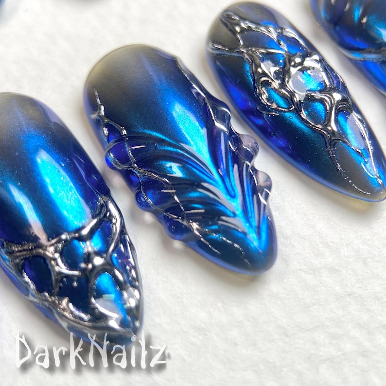 Pu&ograve; includere: Primo piano di nail art blu e argento con un design astratto a spirale. Le unghie sono lunghe e di forma ovale. Il design ricorda una galassia o una nebulosa.