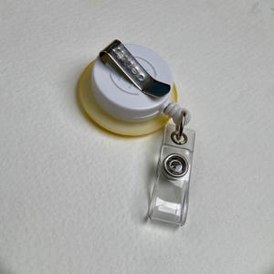 May include: A white and yellow retractable badge reel with a metal clip and a clear plastic badge holder. The reel has a circular design with a metal clip on top and a clear plastic holder attached.