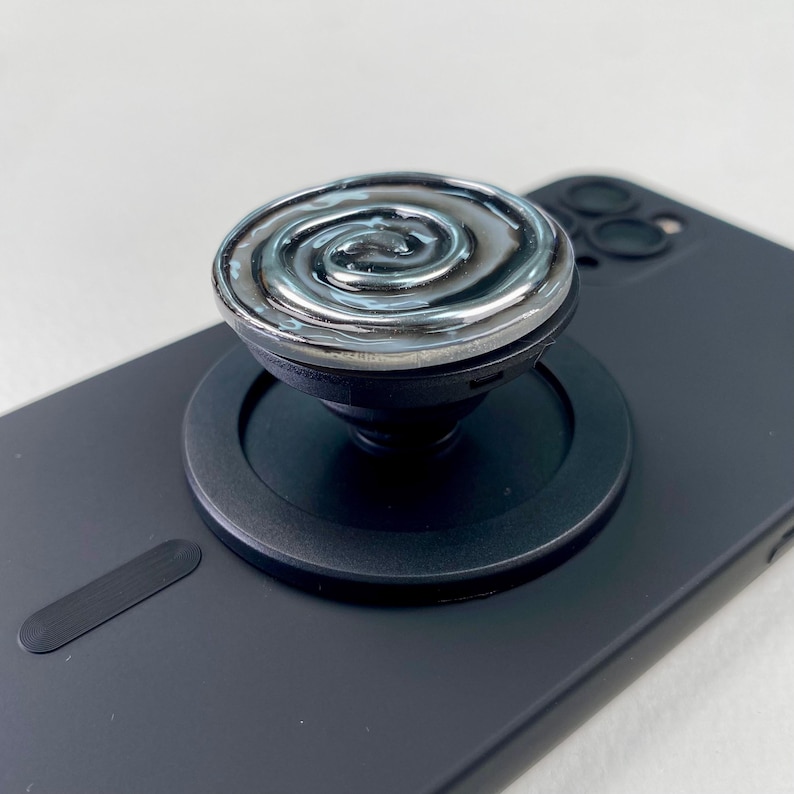 May include: A black phone grip with a circular top featuring a raised, metallic silver swirl pattern. The grip is attached to the back of a matte black smartphone, which has a circular magnetic ring base surrounding the central collapsible stand.