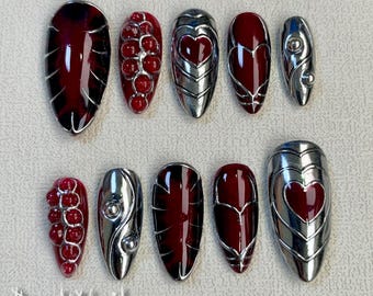 Puede incluir: Conjunto de diez uñas postizas rojas y plateadas con varios diseños, incluyendo corazones, remolinos y un racimo de cuentas rojas. Las uñas están dispuestas en dos filas sobre una superficie blanca. El texto "DarkNailz" es visible en la esquina inferior izquierda de la imagen.