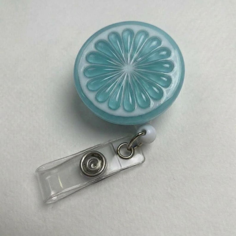 May include: A badge reel with a turquoise and white citrus slice design. The retractable reel has a clear plastic strap and a metal clip. The badge reel is round and has a white border.
