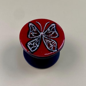 Peut inclure: Un support de téléphone circulaire doté d'une surface rouge brillante avec un emblème de papillon argenté en relief. Le motif du papillon présente des détails en filigrane argenté et de petites perles argentées sur les ailes, sur une base sombre.