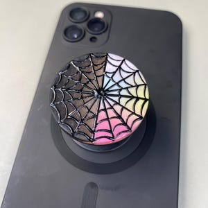 May include: A circular phone grip featuring a raised black spiderweb design over a gradient background that transitions from black on the left to pastel shades of blue, yellow, and pink on the right, attached to a matte black smartphone.