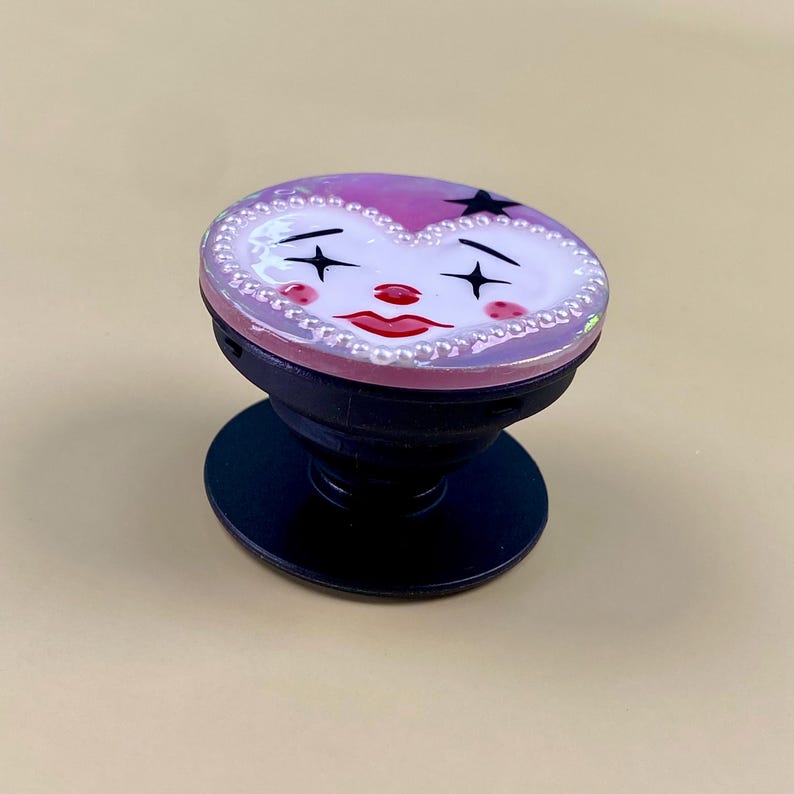Peut inclure: Un support de t&eacute;l&eacute;phone circulaire avec une face en forme de c&oelig;ur au design inspir&eacute; d'un clown. Le visage pr&eacute;sente une peau blanche, des l&egrave;vres rouges, des joues roses et des yeux en forme d'&eacute;toile noire, bord&eacute; de perles blanches.