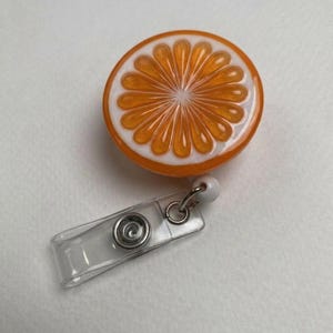 May include: A round badge reel featuring a vibrant orange slice design with white accents. The reel has a clear plastic strap and a metal clip for attaching to clothing or accessories.
