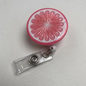May include: A round retractable badge reel featuring a pink grapefruit slice design with white and pink details. The reel has a clear plastic strap and a metal clip for attaching to clothing or accessories.