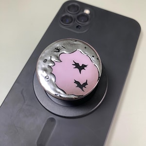 May include: A circular phone grip featuring a metallic silver crescent moon design against a soft pink background. The design includes two small black bat silhouettes flying within the moon's curve. The grip is attached to a matte black smartphone.