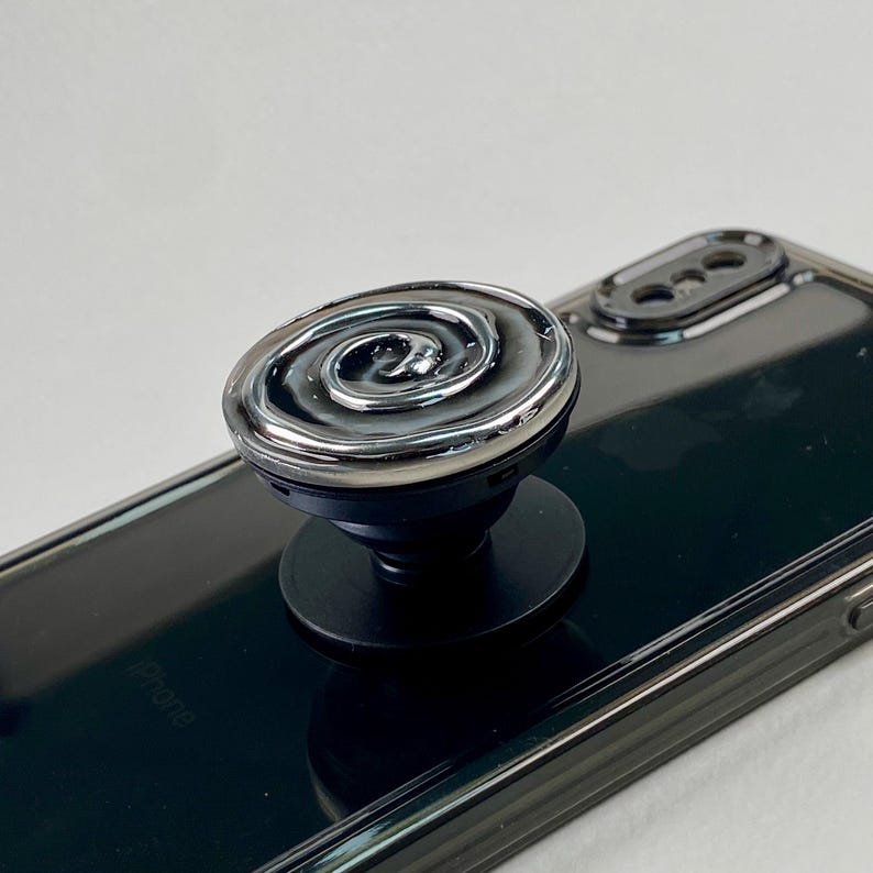 May include: A black phone grip with a circular top featuring a raised, metallic silver spiral design. The accessory is attached to the back of a black smartphone, which has a visible camera lens array and a glossy, reflective surface finish.