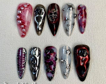 May include: Ten long, pointed false nails with different designs.  Some nails have a red base with black and silver accents, others have a silver base with black accents, and one has a pink base with white swirls and silver pearls.  The nails are arranged in two rows of five.