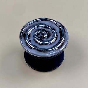 May include: A circular phone grip featuring a glossy, swirling pattern of black and white resin. The top surface displays a raised, metallic-toned spiral design that contrasts with the smooth, marbled background. It has a solid black base.