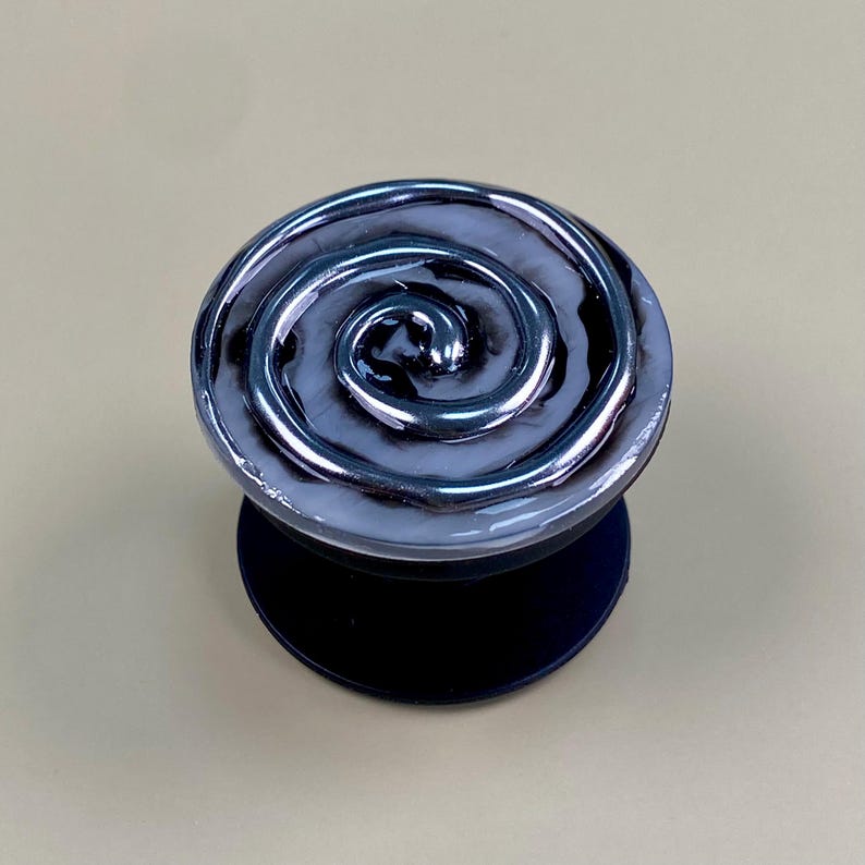 May include: A circular phone grip featuring a glossy, swirling pattern of black and white resin. The top surface displays a raised, metallic-toned spiral design that contrasts with the smooth, marbled background. It has a solid black base.