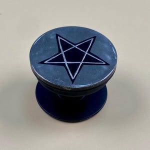 May include: A circular phone grip featuring a black pentagram design centered on a dark, textured metallic surface. The device has a collapsible black plastic base and a smooth, rounded top edge, providing a functional accessory for mobile devices.