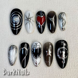 Puede incluir: Diez puntas de uñas ovaladas en blanco y negro con diseños plateados y negros. Los diseños incluyen un corazón, una telaraña, una luna creciente, una estrella y una cruz.