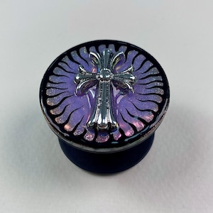 May include: A circular phone grip featuring a metallic silver cross emblem centered on a purple, glittery, sunburst-patterned background. The design is encased in a glossy, dark-rimmed finish, providing a reflective surface for the cross.
