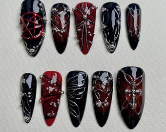 May include: A set of ten long, pointed, press-on nails. Each nail features a black and red gradient design with silver embellishments. Some nails have pentagrams, crosses, or intricate silver patterns.