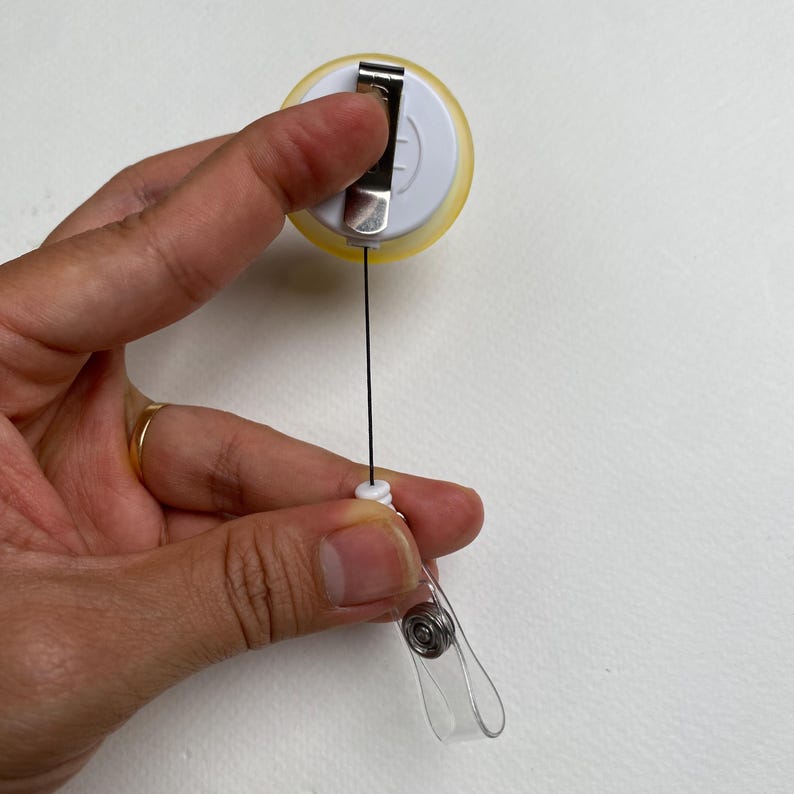 May include: A yellow and white retractable badge reel with a metal clip. The reel has a black cord and a clear plastic clip at the end. The reel is held in a person's hand.