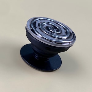 May include: A black collapsible phone grip featuring a circular top with a raised, metallic silver spiral design encased in a clear, glossy resin finish. The base is a solid black plastic disc with an accordion-style stem for adjustable height.