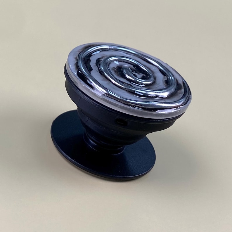 May include: A black collapsible phone grip featuring a circular top with a raised, metallic silver spiral design encased in a clear, glossy resin finish. The base is a solid black plastic disc with an accordion-style stem for adjustable height.