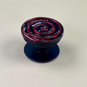 Könnte beinhalten: Ein runder Handyhalter mit einer glänzend schwarzen Basis und einer Oberseite, die mit einem leuchtend roten Spiralmuster auf tiefschwarzem Grund verziert ist. Das Zubehör verfügt über einen faltbaren Akkordeon-Stiel und eine flache Montageplatte.