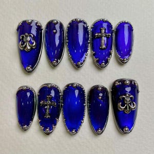 May include: Set of ten artificial nails in a deep blue hue, each with a silver-colored trim. Some nails feature a silver cross or decorative emblem. The nails have a glossy finish and are arranged on a light surface.