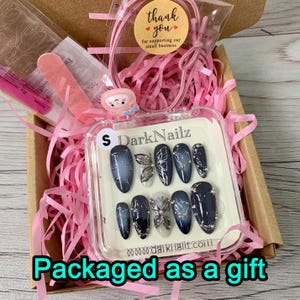 Pu&ograve; includere: Una scatola di plastica trasparente riempita con carta crespa rosa e un set di unghie finte nere e argentate. La scatola &egrave; etichettata "DarkNailz" e ha una piccola "S" in alto. Le unghie hanno un design a farfalla e sono confezionate come regalo.