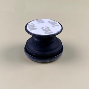 May include: A black collapsible phone grip featuring a circular top with a white adhesive backing. The white surface displays the repeating text 3M in a grey font. The device has a tiered, accordion-style base made of black plastic material.