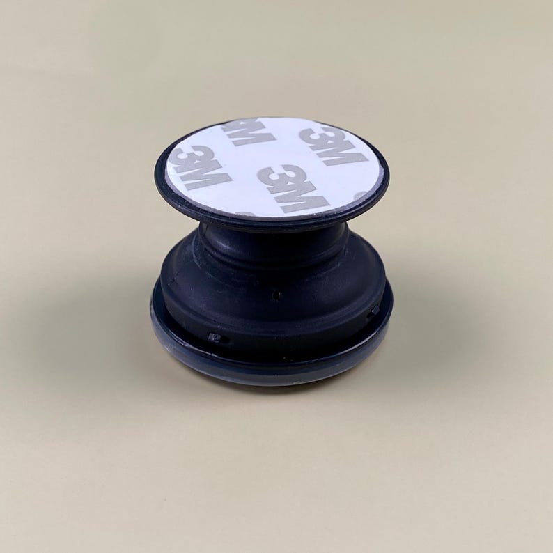 May include: A black collapsible phone grip featuring a circular top with a white adhesive backing. The white surface displays the repeating text 3M in a grey font. The device has a tiered, accordion-style base made of black plastic material.