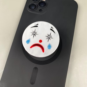 Puede incluir: Un soporte circular blanco para teléfono con un diseño de cara de payaso triste. La cara tiene cejas negras arqueadas, dos ojos en forma de estrella con pequeños cristales incrustados, una nariz roja brillante, una boca roja triste y dos lágrimas azules.