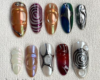 May include: A collection of ten artificial nails with various designs. The nails feature colors like iridescent orange, purple, silver, and pearl. Some nails have swirl patterns, while others have 3D embellishments and metallic accents. The text "DarkNailz" is visible at the bottom.
