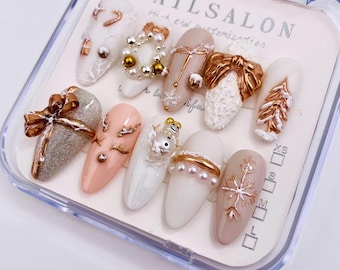 Puede incluir: Un conjunto de diez uñas artificiales blancas y beige con varios diseños de temática invernal. Los diseños incluyen un muñeco de nieve, un copo de nieve, un árbol de Navidad, un regalo y un reno. Las uñas se muestran en una caja de plástico transparente con el texto "NAILS SALON High-end customization" impreso en la parte superior.