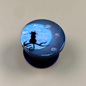 Puede incluir: Un soporte circular para teléfono con fondo azul oscuro y una gran luna azul claro. Una silueta negra de un gato está sentada sobre una rama de árbol frente a la luna. La superficie tiene varias gotas circulares pequeñas, claras y en relieve.