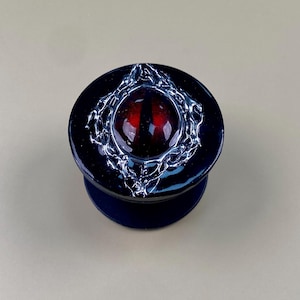 Peut inclure: Un support de téléphone circulaire avec une base noire brillante et un motif central en relief représentant un œil rouge à pupille noire verticale. L'œil est entouré d'un cadre texturé argenté métallique, rappelant une toile ou des lianes.