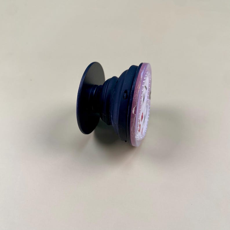 Peut inclure: Un support de t&eacute;l&eacute;phone r&eacute;tractable avec une base circulaire bleu fonc&eacute; et une tige en accord&eacute;on. La surface sup&eacute;rieure est un disque plat avec une bordure rose clair et un motif central compos&eacute; d'&eacute;l&eacute;ments blancs et rouges sur fond clair.