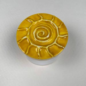 May include: A circular lid featuring a raised, glossy yellow sun design with a central spiral and radiating rays outlined in white. The lid has a smooth, reflective finish and sits atop a plain, white cylindrical base container.