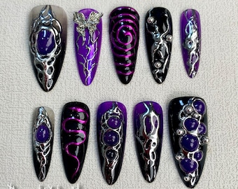 Könnte beinhalten: Satz mit zehn lila und schwarzen Nagelspitzen mit silbernen Akzenten. Die Spitzen haben verschiedene Designs, darunter Spiralen, Schmetterlinge und Edelsteine.