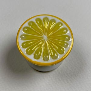 Peut inclure: Un bouton de meuble circulaire arborant un motif de tranche d'agrume jaune et blanc. La surface supérieure présente un motif radial de segments jaunes sur fond blanc, entouré d'une bordure jaune unie sur une base cylindrique blanche.