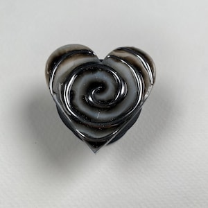 May include: A heart-shaped decorative knob featuring a glossy, swirling pattern of translucent gray, white, and deep black tones. The surface has a smooth, reflective finish with a raised spiral design that radiates from the center of the heart.