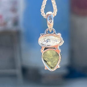 May include: A silver necklace with a pendant featuring a pear-shaped green gemstone and a smaller round clear gemstone. The pendant has a silver tag with the inscription "925".