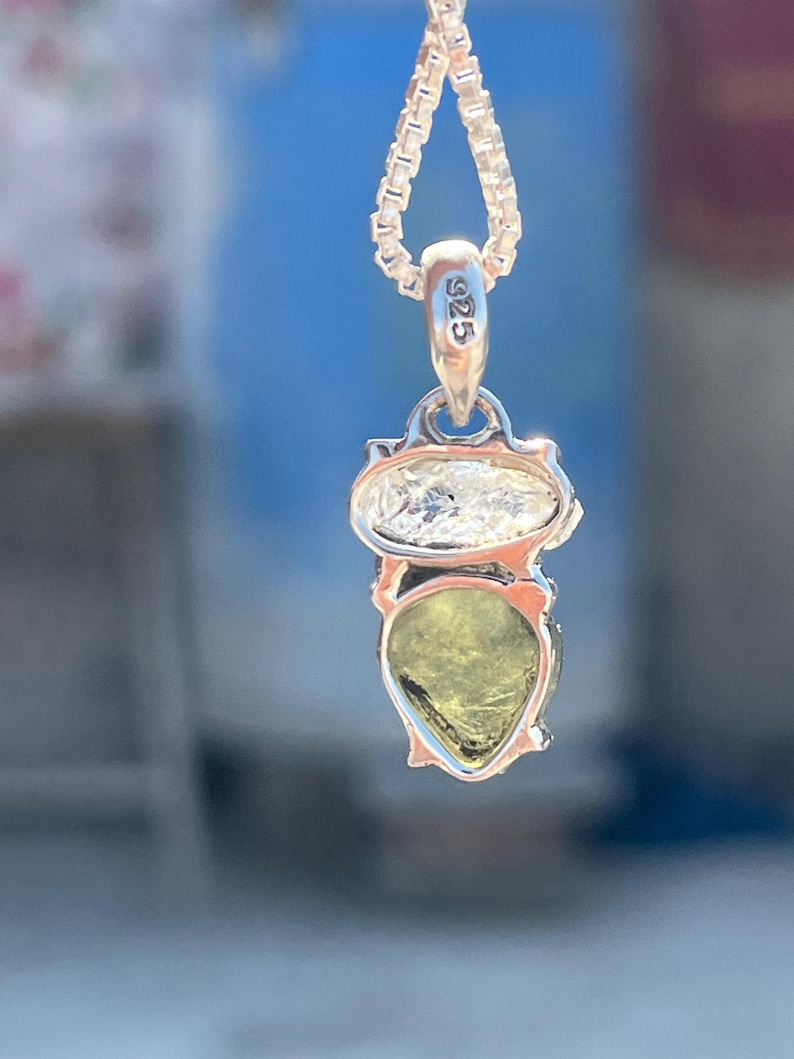 May include: A silver necklace with a pendant featuring a pear-shaped green gemstone and a smaller round clear gemstone. The pendant has a silver tag with the inscription "925".