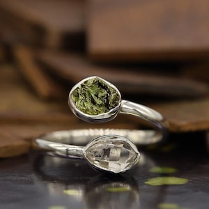 May include: A silver ring featuring two oval-shaped gemstones. One gemstone is a vibrant green, while the other is clear. The ring's design allows the gemstones to sit on top of the finger, creating a unique and eye-catching look.