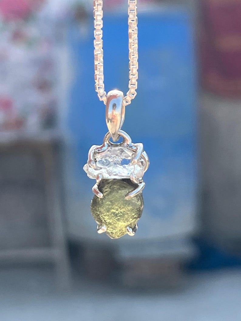 May include: A silver chain necklace with a pendant featuring a green gemstone set in a silver bezel. The gemstone is surrounded by a silver halo.