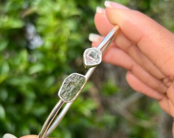 May include: A silver bangle bracelet with two raw, faceted, green and clear gemstones. The bracelet is open and has a simple design.