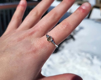 Op de afbeelding: Een zilveren ring met een kleine, ovale, bruine edelsteen in het midden. De ring zit aan een vinger.